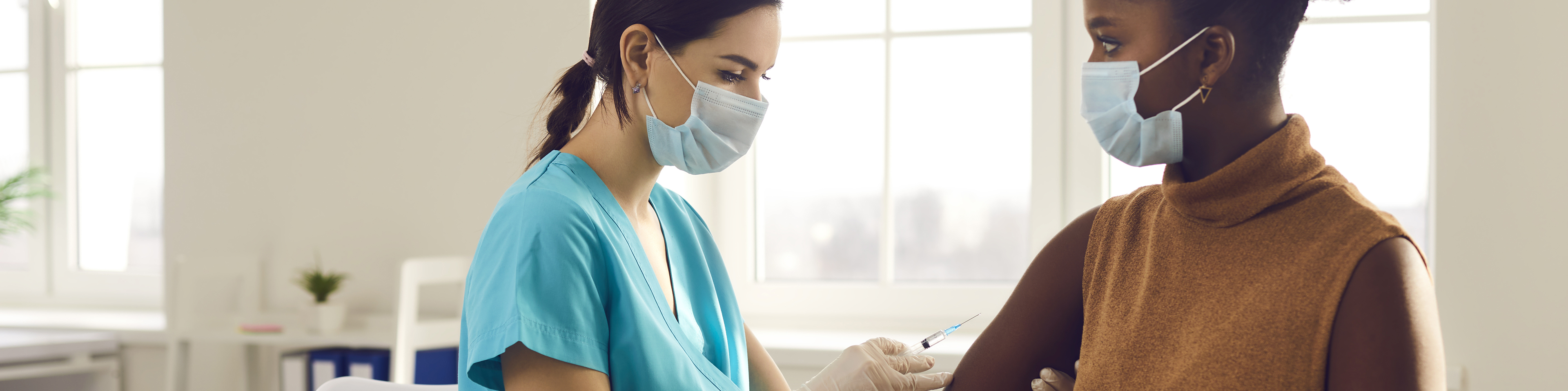 Young African American woman in medical face mask sitting at doctor's office and getting a shot in her arm, nurse in medical face mask holding syringe and giving female patient modern covid 19 vaccine injection