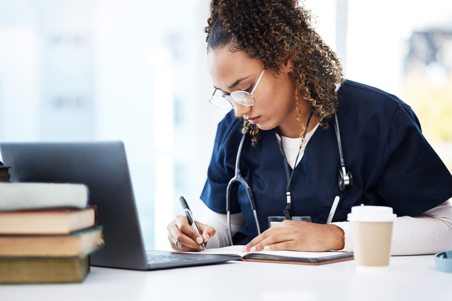 Medical professional, wearing blue scrubs, uses laptop to conduct research; a cup of coffee is in the foreground