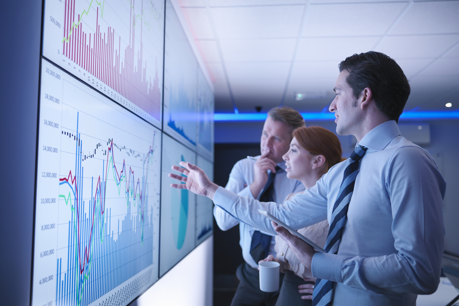 Three business people discuss graphs on screen in meeting room