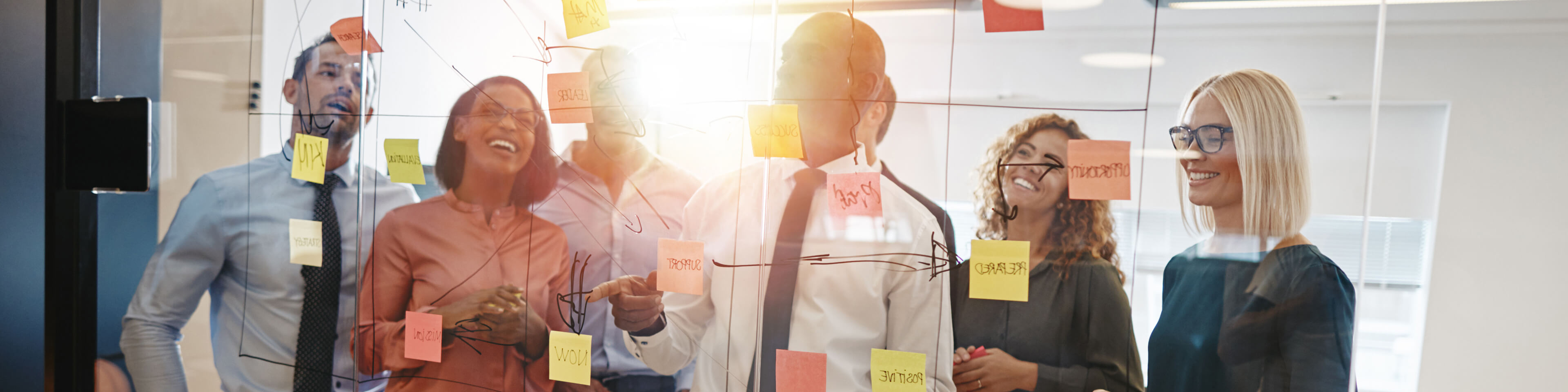 Group of men and women standing at a whiteboard, looking at a flowchart with sticky notes on it. the people are smiling.