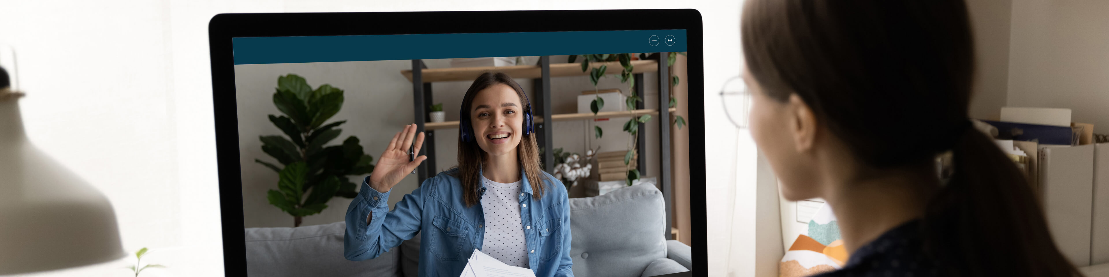 woman wearing glasses looking at a laptop screen, where another woman sits on a couch and waves, smiling.