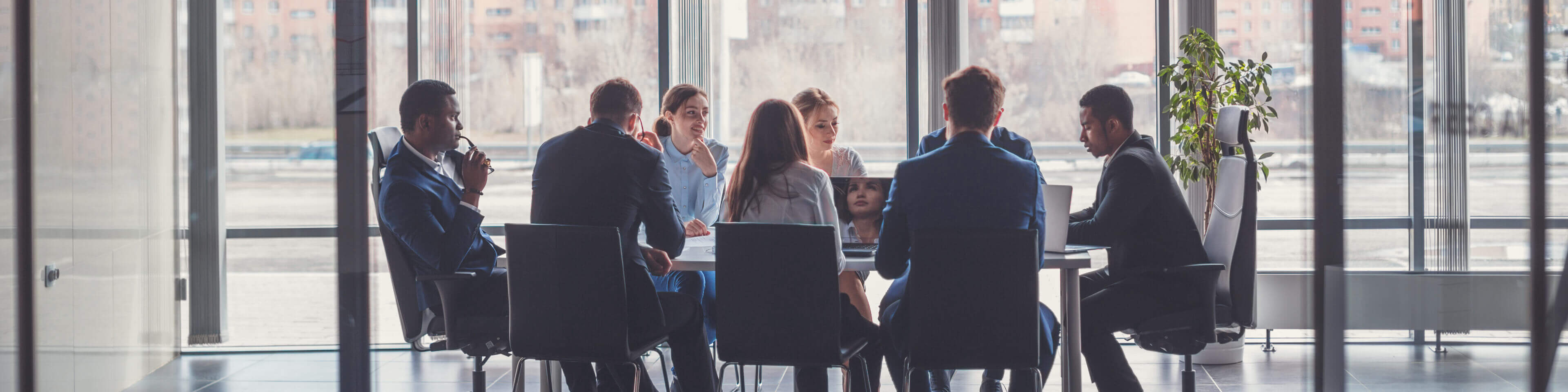 Group of men and women in business attire sitting at a conference table, talking and looking at laptops