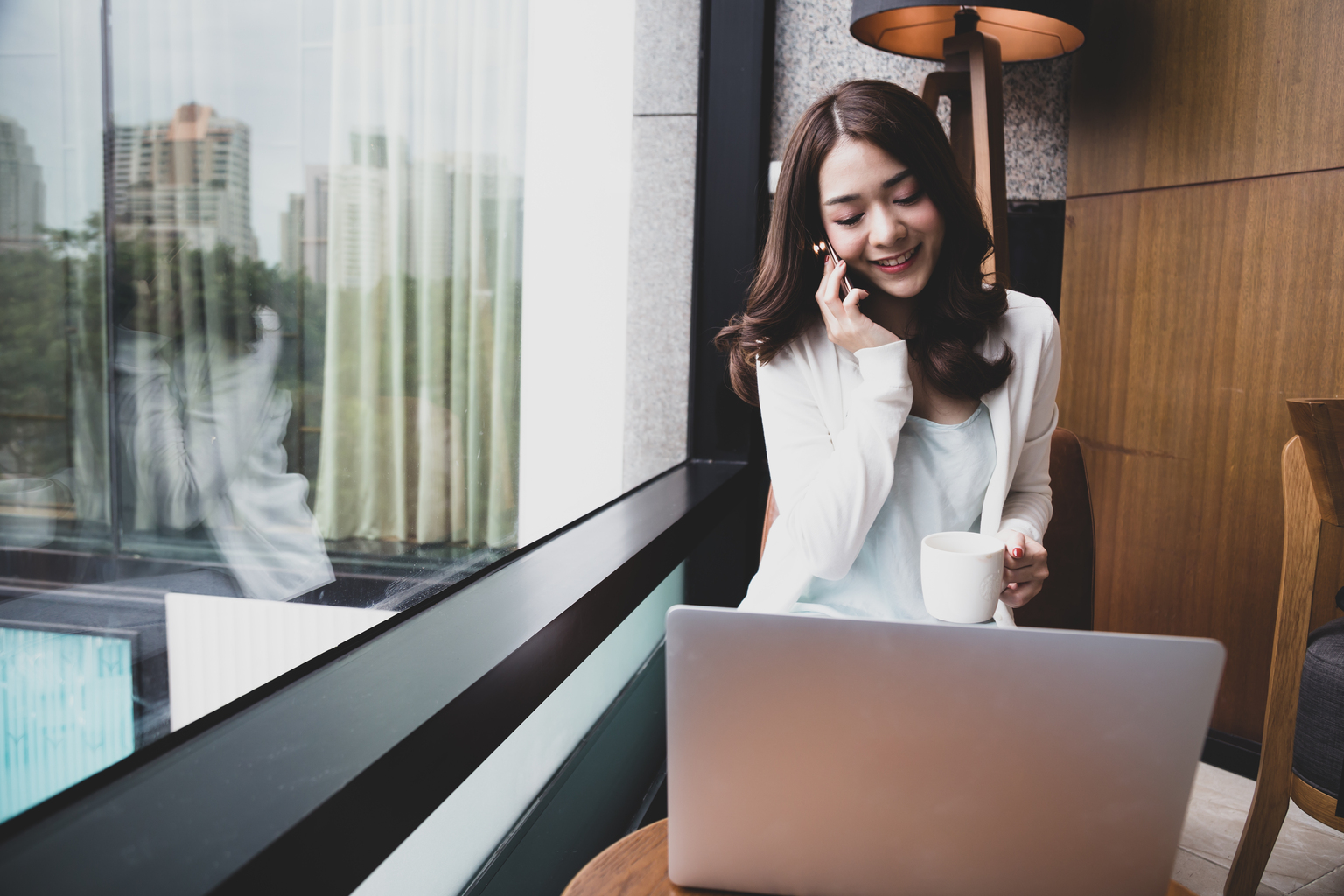 Young Asian woman using phone and laptop with coffee next to window