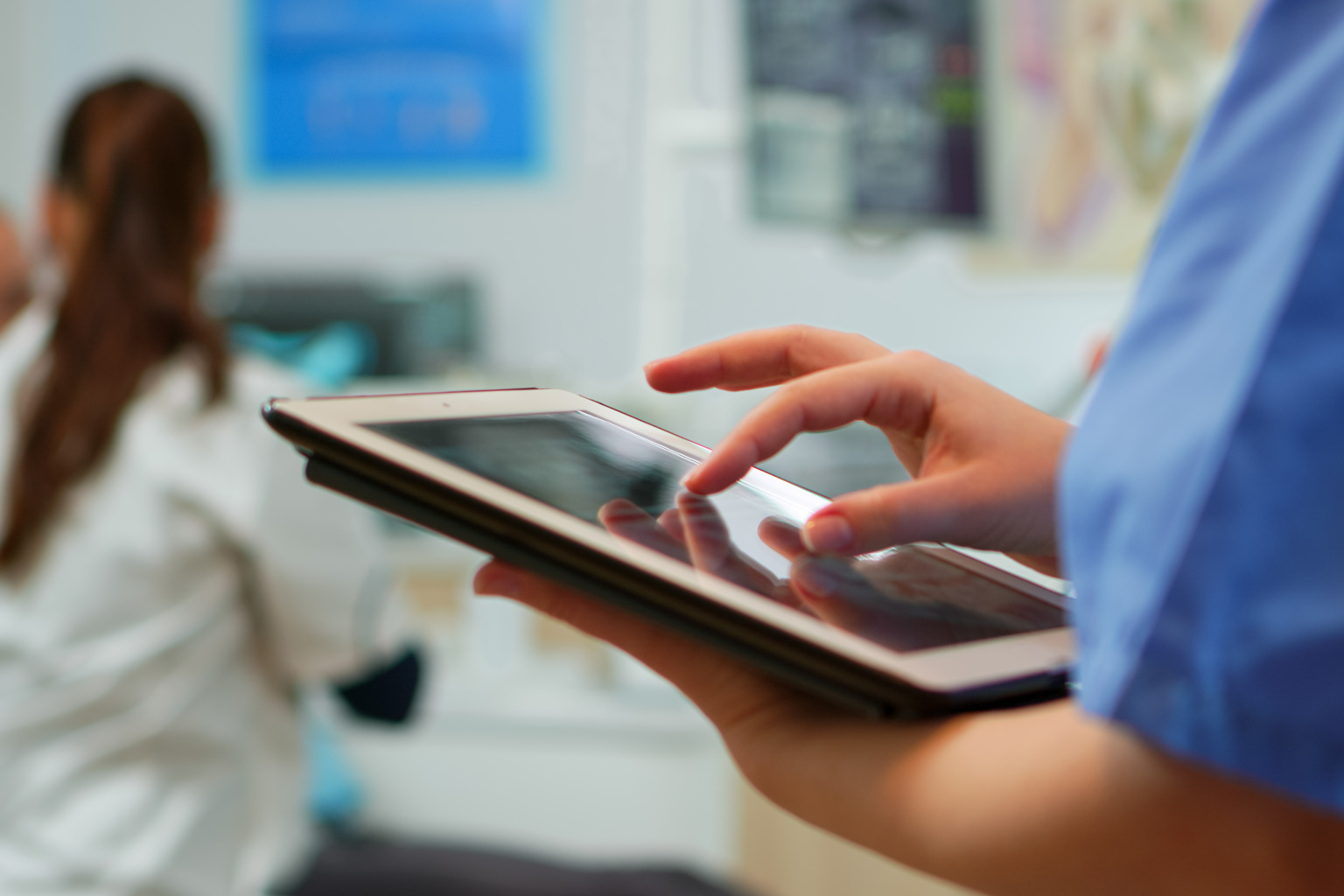 Close up of nurse holding and typing on tablet standing in stomatologic clinic, while doctor is working with patient in background