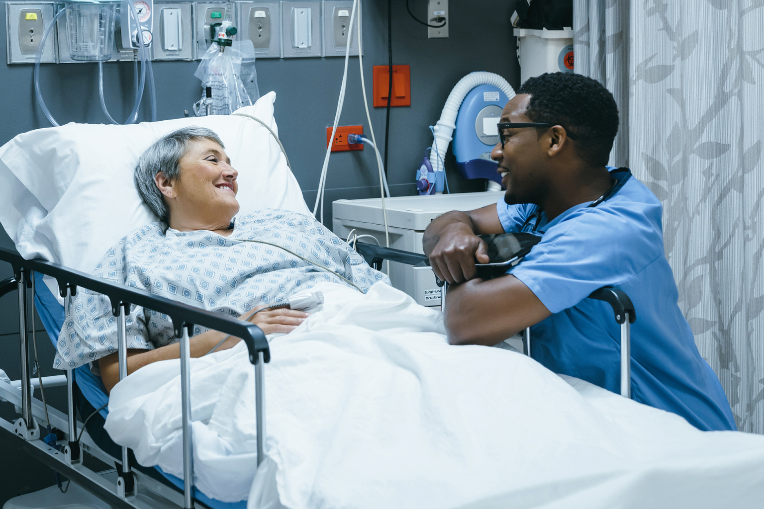 African male nurse talking to senior white female patient in hospital bed.
