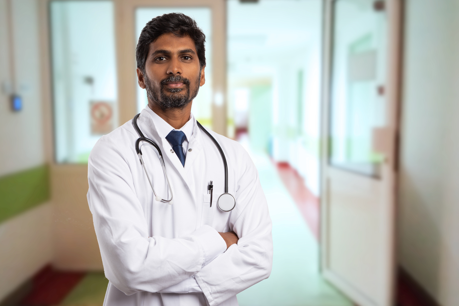 Portrait of male indian doctor with serious expression and crossed arms wearing white coat having open door on clinic corridor as background