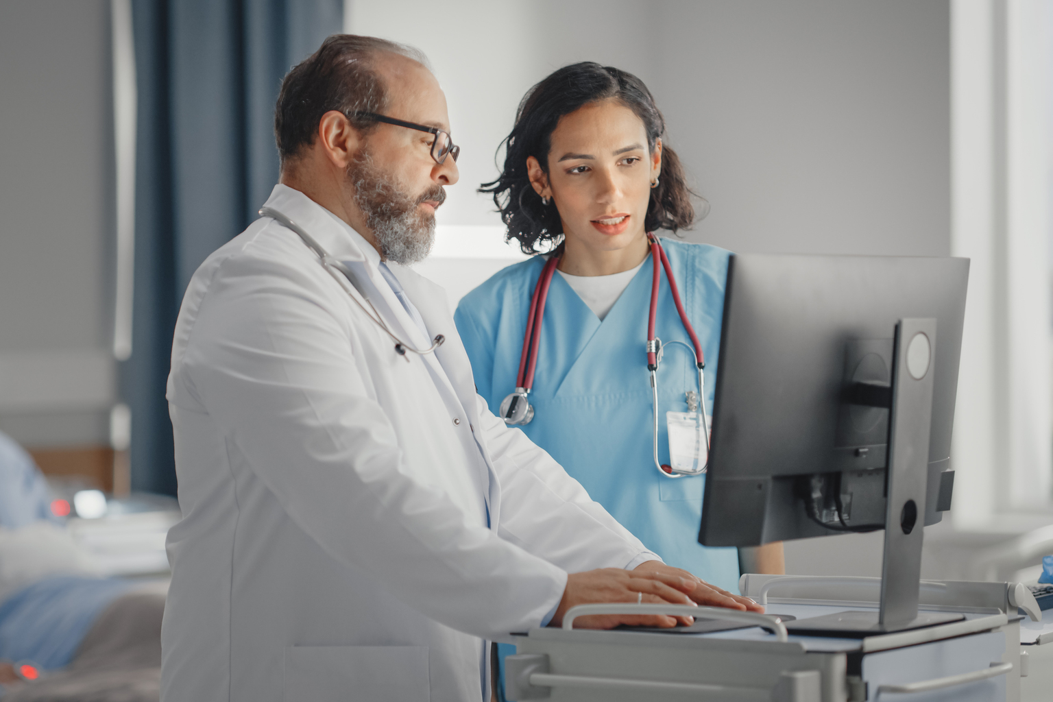 Doctor talks with head nurse at computer with patient in hospital bed in the background