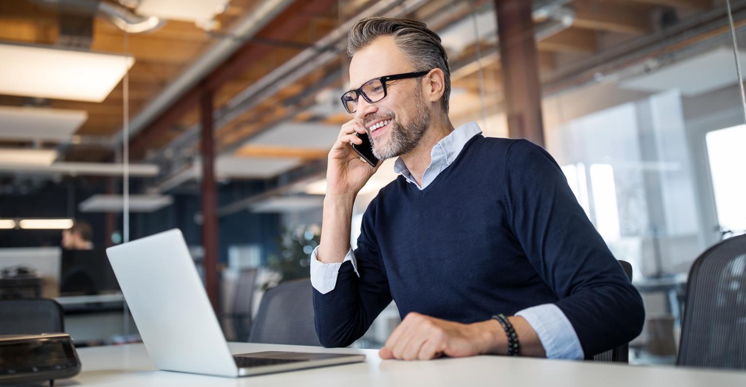 Mid adult business professional sitting at table with laptop and talking on mobile phone. Businessman working in office.