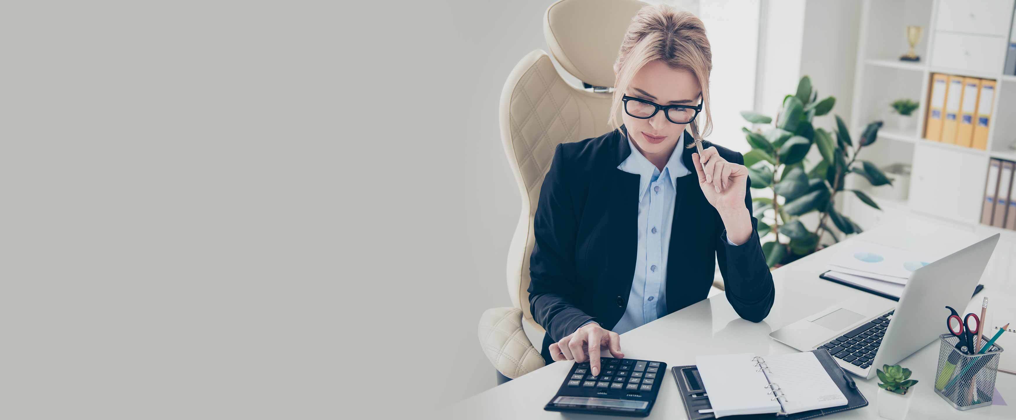 Portrait of busy concentrated woman in eyewear counting on calculator with thoughtful expression wearing shirt jacket sitting in modern workplace having computer supplies on table