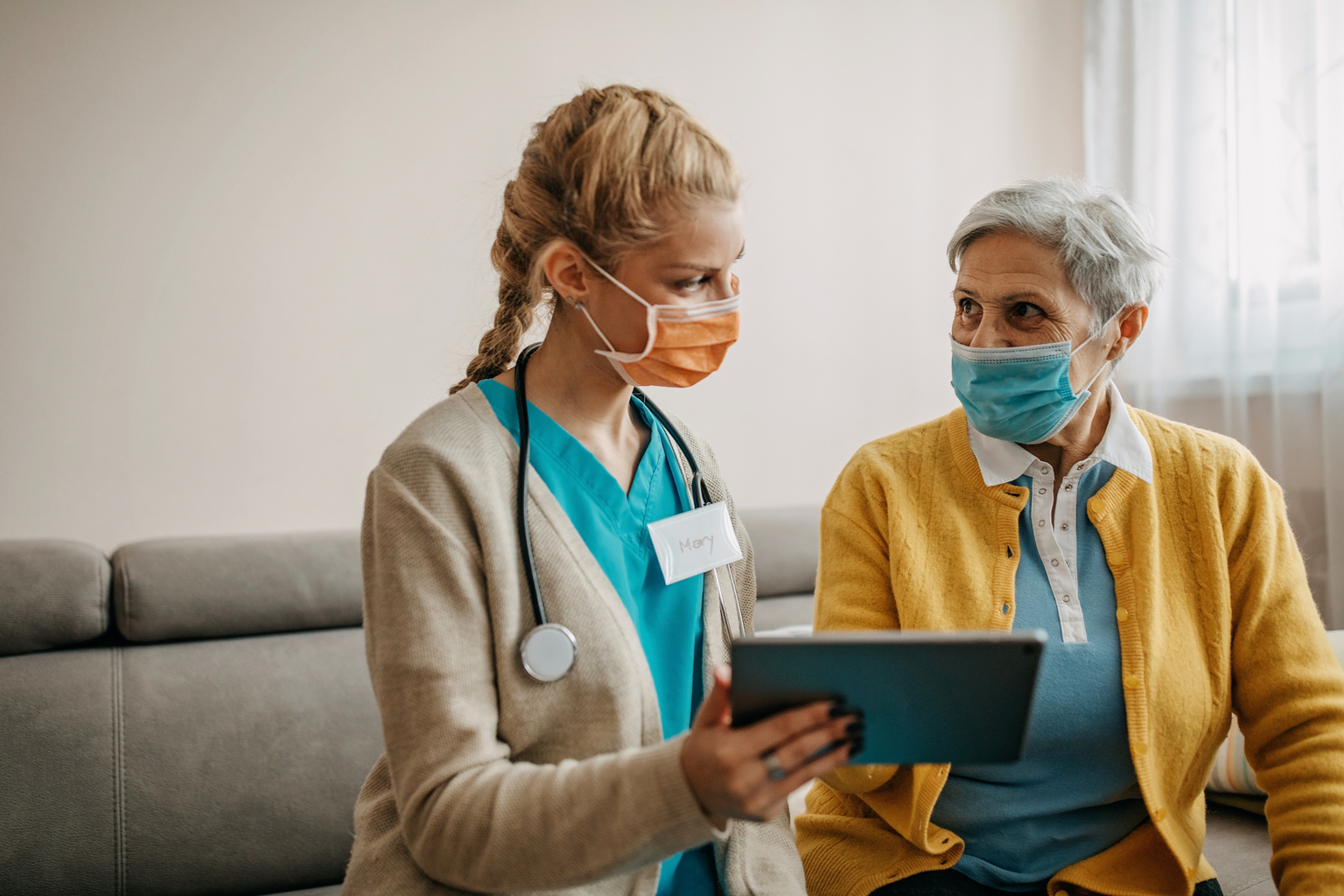 a female nurse with blue scrubs and light brown cardigan talking to patient and using laptp