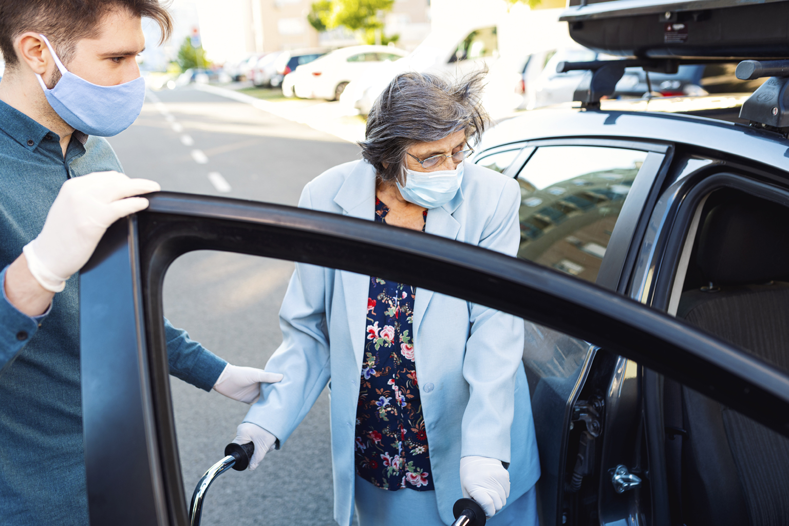 Young man helping older woman with walker get into car, both wearing masks