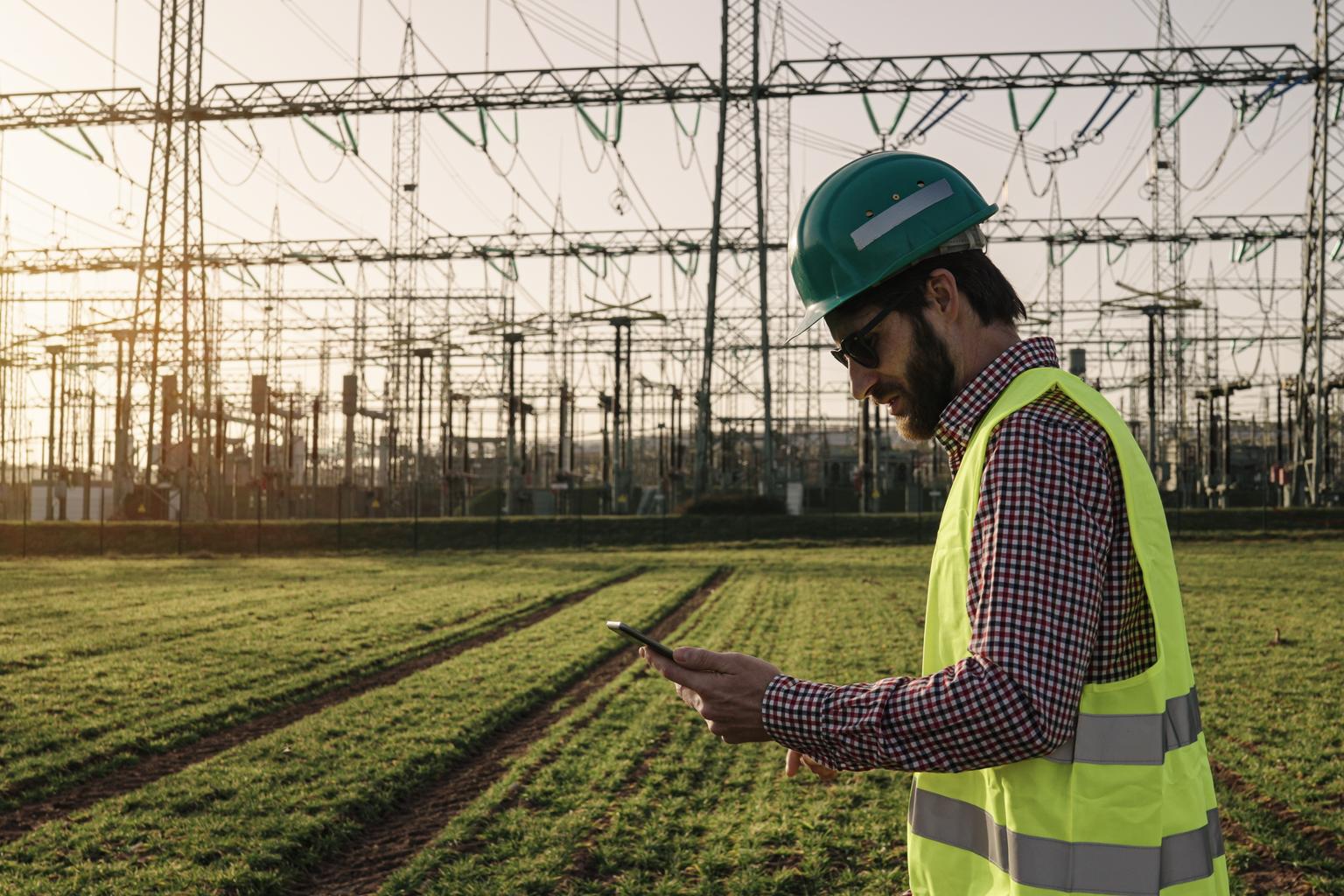 Electrical engineer wearing a helmet and safety vest working with tablet near high voltage electrical lines power station during sunset