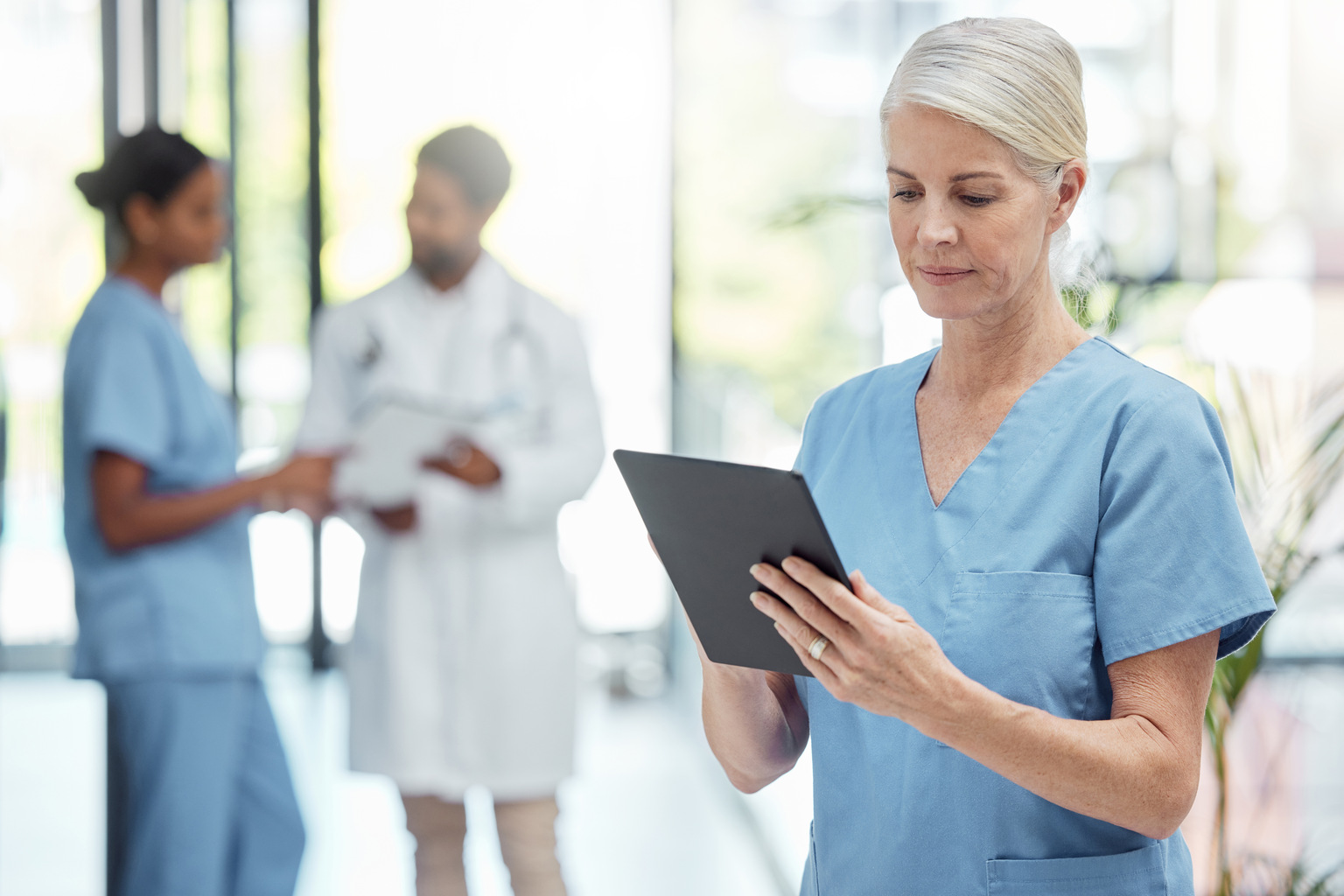 Experienced nurse reviewing patient chart on tablet; two colleagues standing to her side