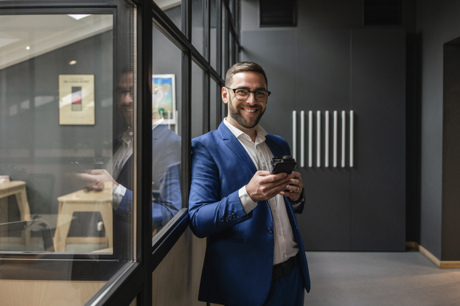 Smiling male professional holding mobile phone and coffee cup while standing by glass wall in office corridor