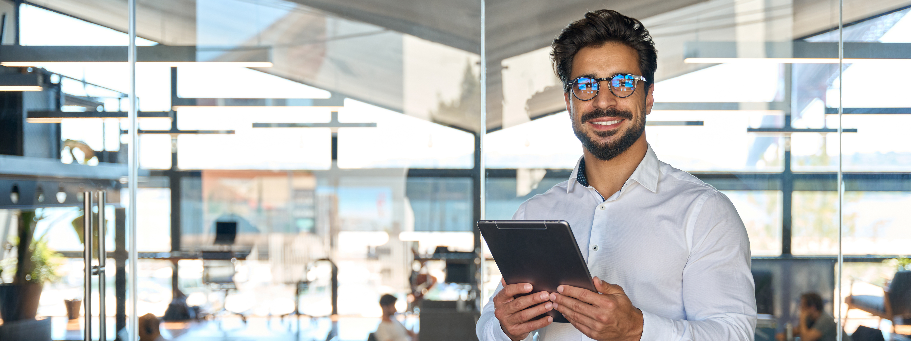 Smiling young business man entrepreneur using tablet standing in office at work. Happy male professional executive ceo manager holding tab computer looking away and thinking of tech data.