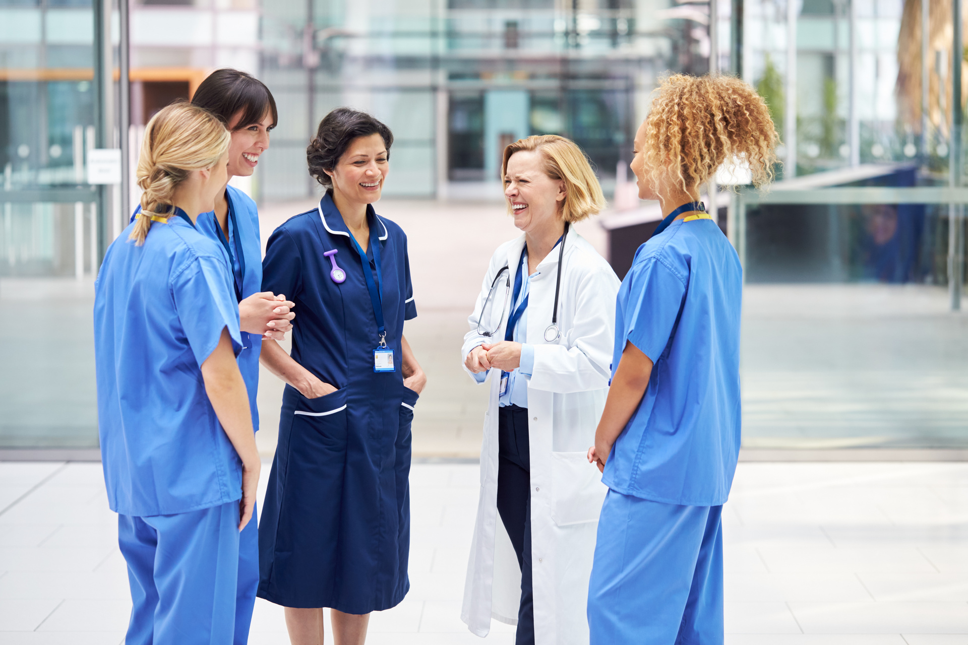 Female Medical Team Having Informal Meeting Standing In Lobby Of Modern Hospital Building