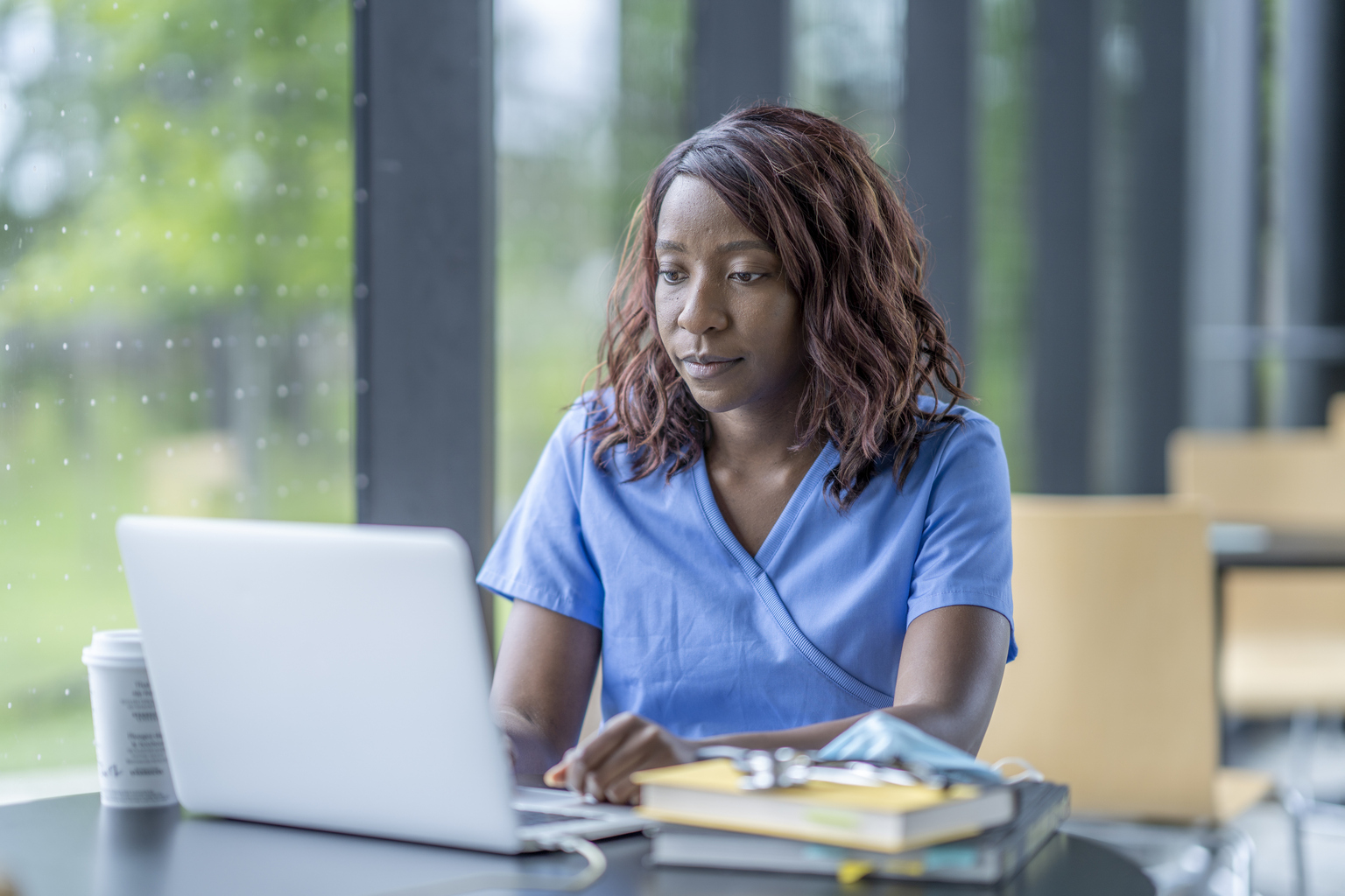 A nurse wearing blue scrubs, is studying at a table scatted with books and a cup of coffee