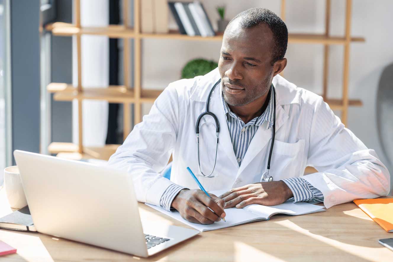 Black doctor looking at computer and taking notes while sitting at workplace