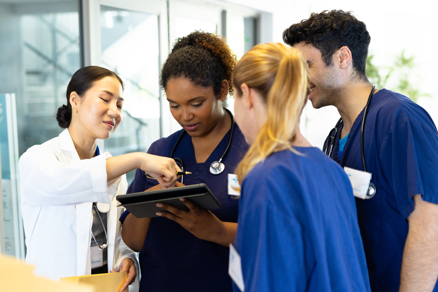 Four happy diverse healthcare colleagues looking at tablet together in hospital corridor