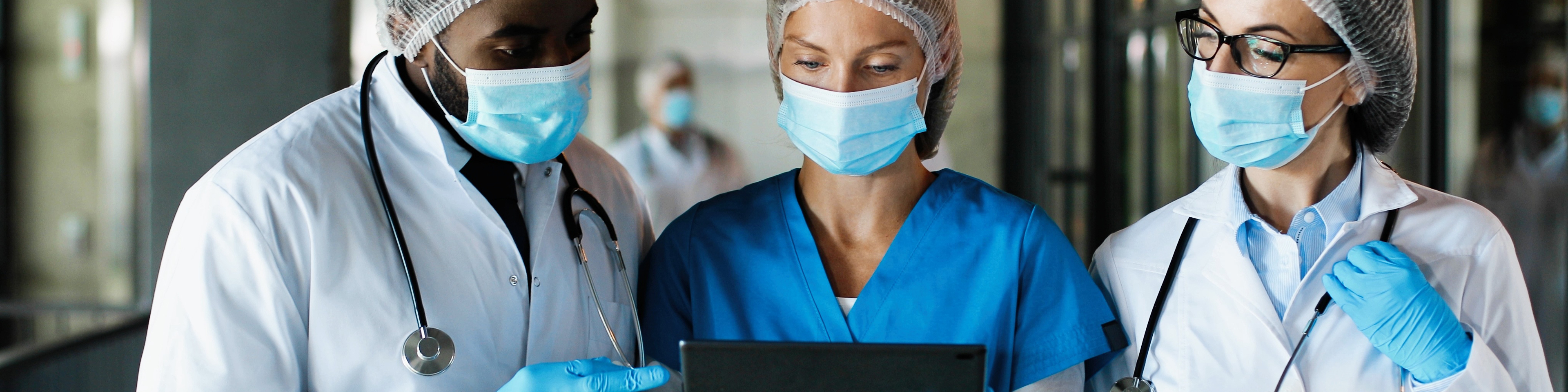 Mixed races male and females healthcare workers in hats medical masks and gloves walking in hospital and talking using tablet device