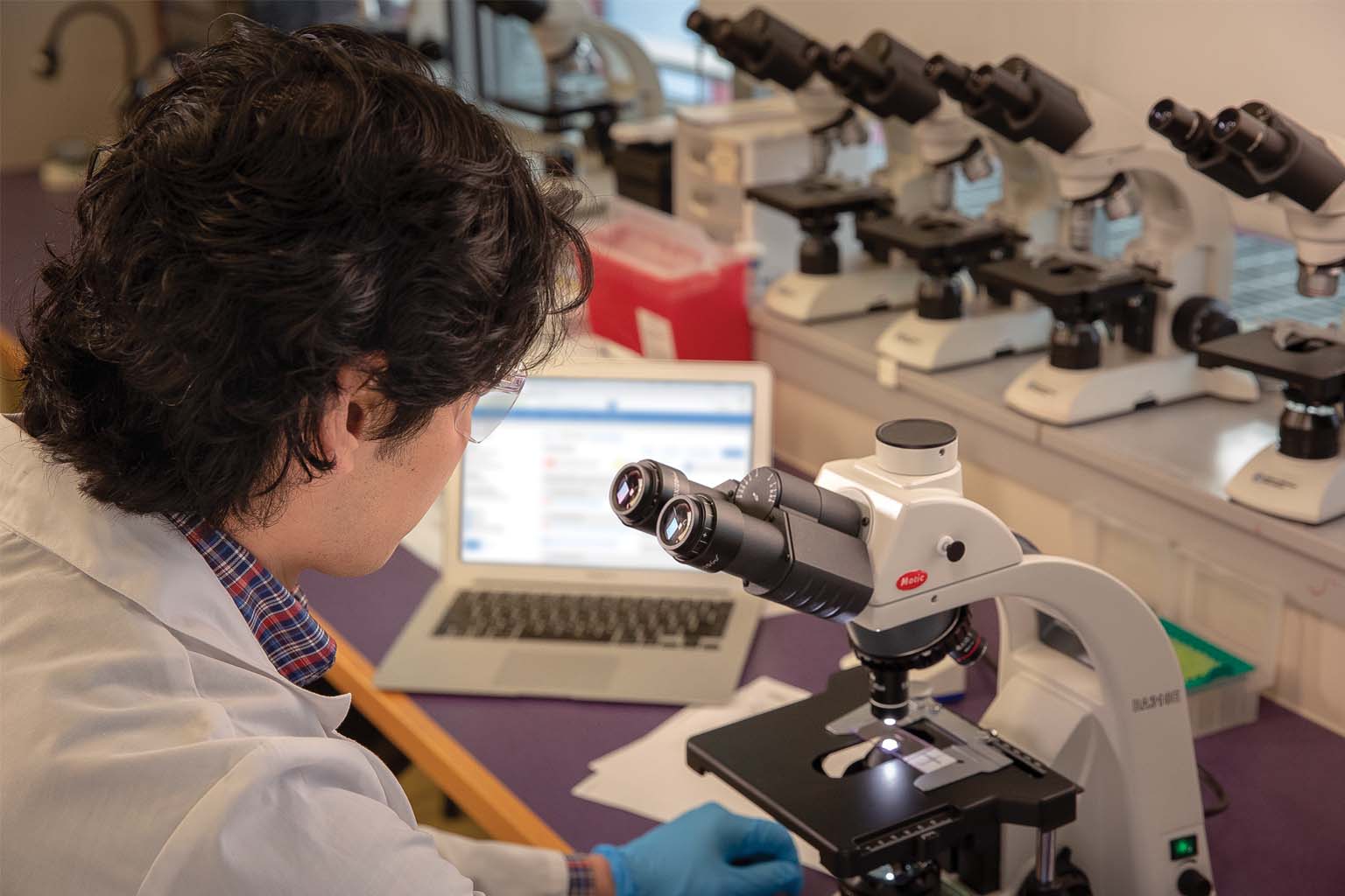 technician in lab with microscope and laptop