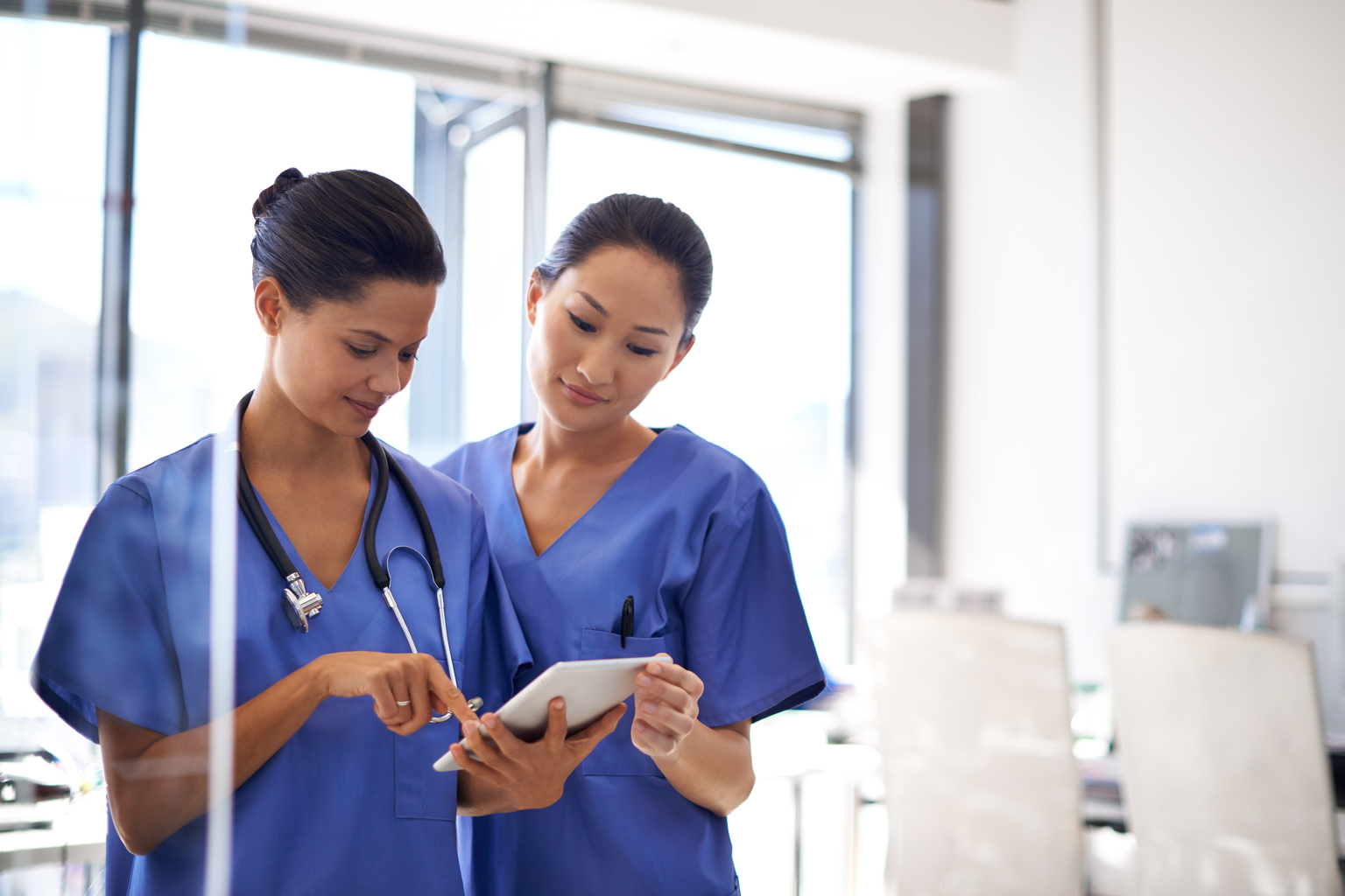 Two female nurses, white and Asian, using a tablet to review medical records.