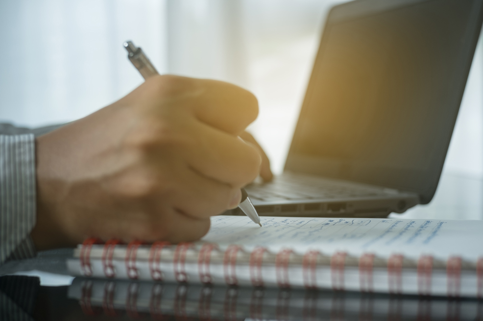 Close up on hand writing in notebook with laptop on the table behind