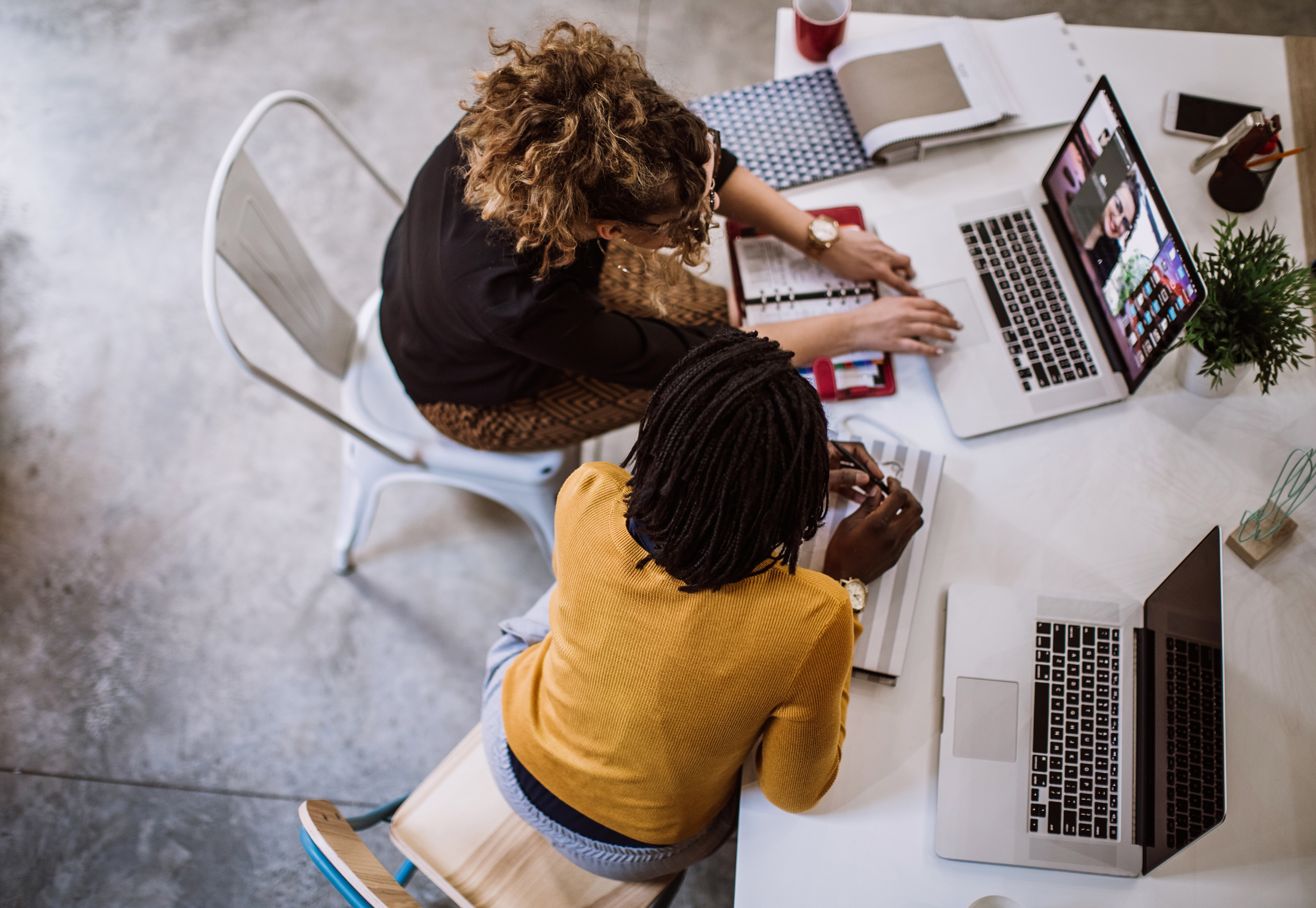 Above photography shot of two, 2 women sitting at office space, one using facetime, video on macbook, laptop, red planner, Q3 2021, TAA NA US - Preparer