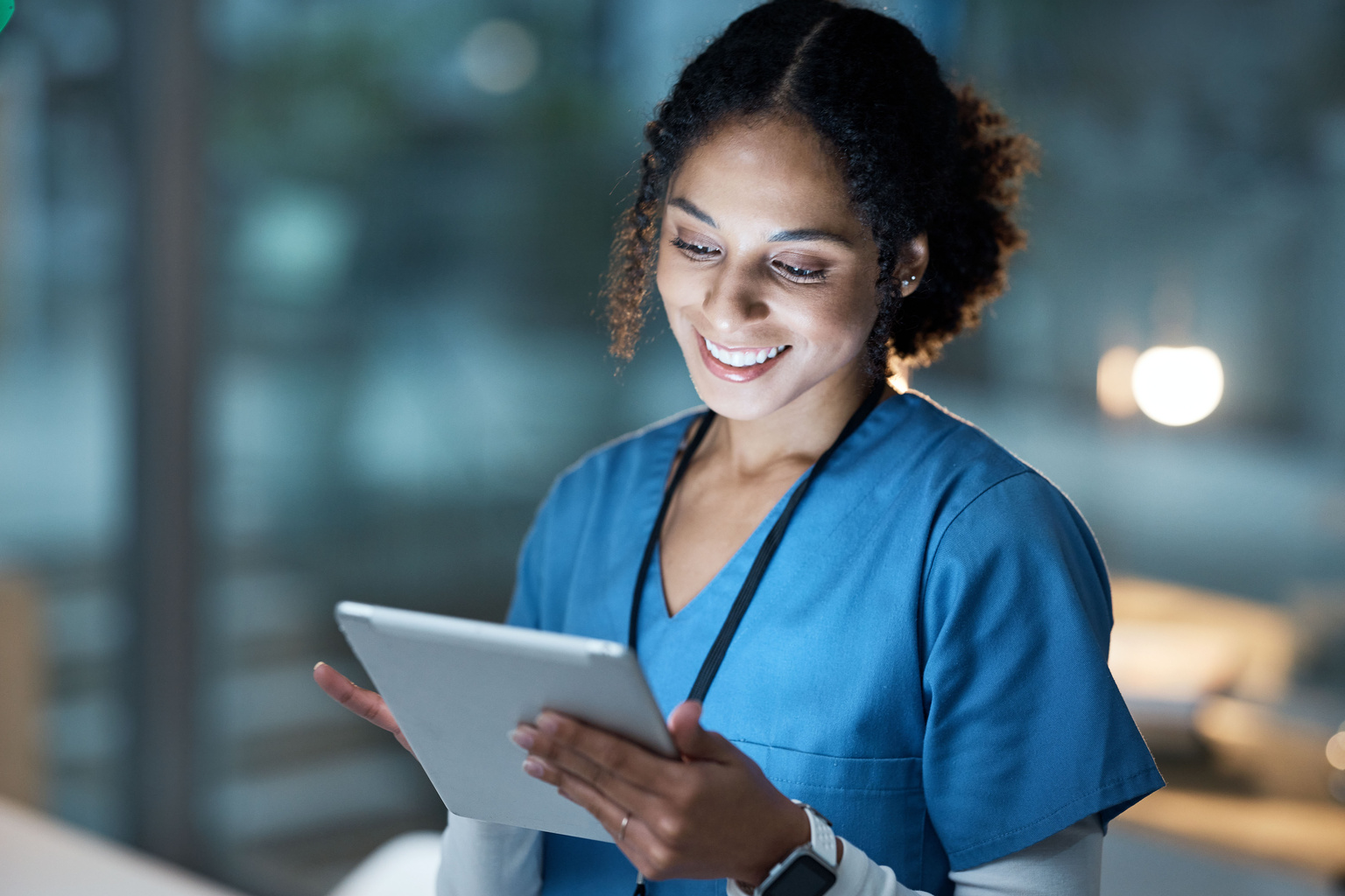 Female nurse wearing blue scubs in a dark room reading information from a tablet