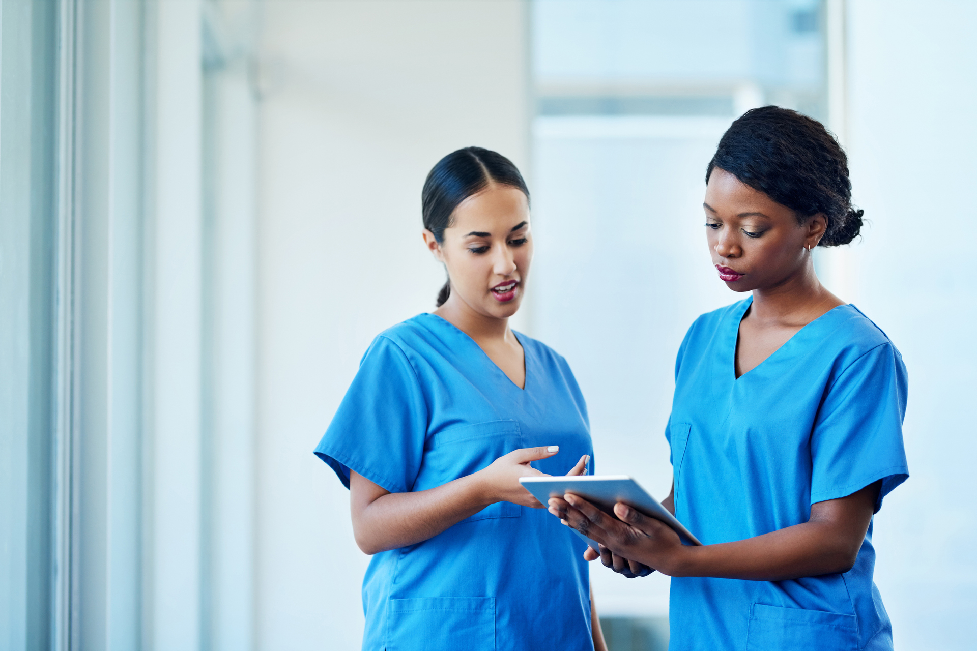 Two nurses wearing blue scrubs discuss patient record on tablet