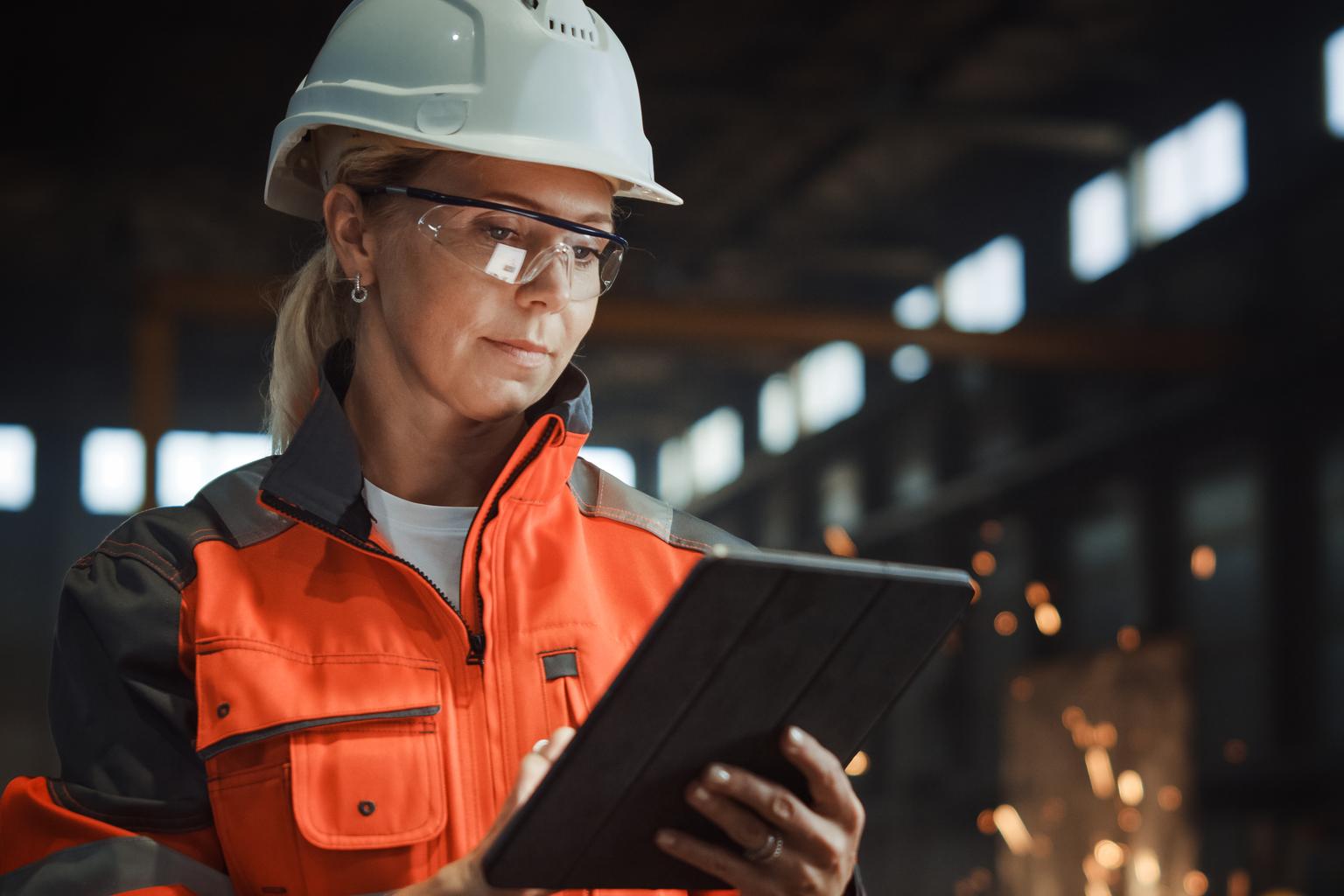 Professional Heavy Industry Engineer Worker Wearing Safety Uniform and Hard Hat Uses Tablet Computer. Serious Successful Female Industrial Specialist Standing in a Metal Manufacture Warehouse.