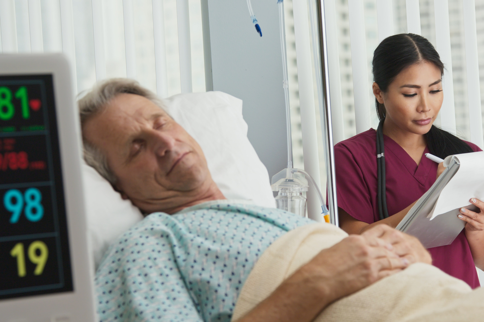 Medical professional taking notes on chart with senior caucasian male patient recovering in hospital bed