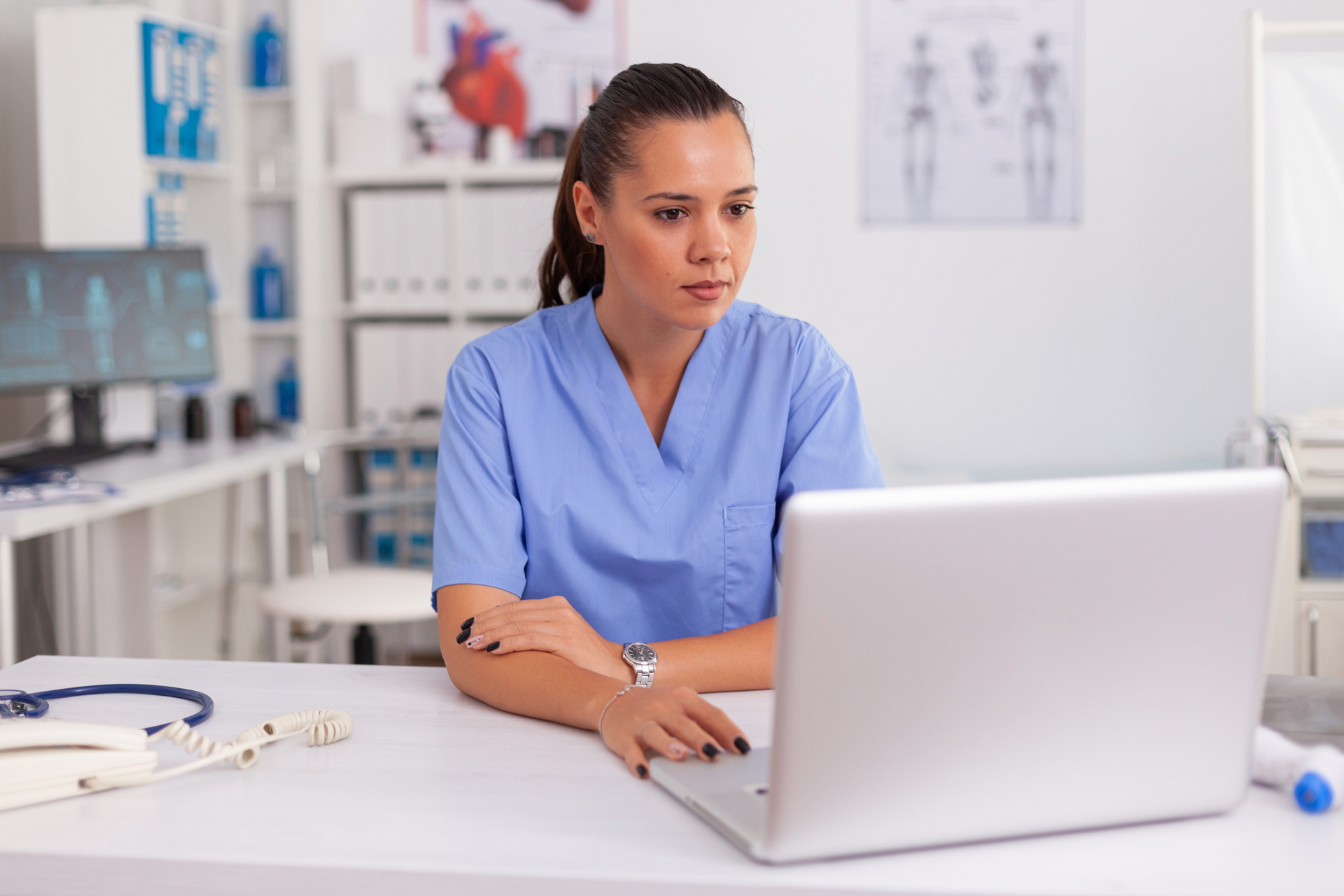 A nurse working on a laptop in a medical office with anatomy charts hanging on the wall in the background