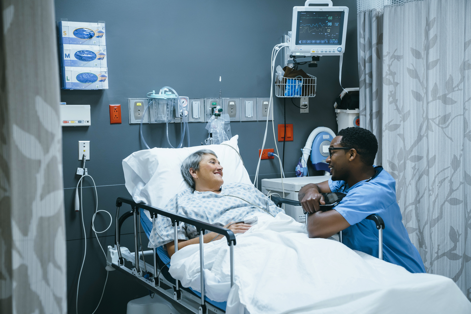 African male nurse talking to senior white female patient in hospital bed.