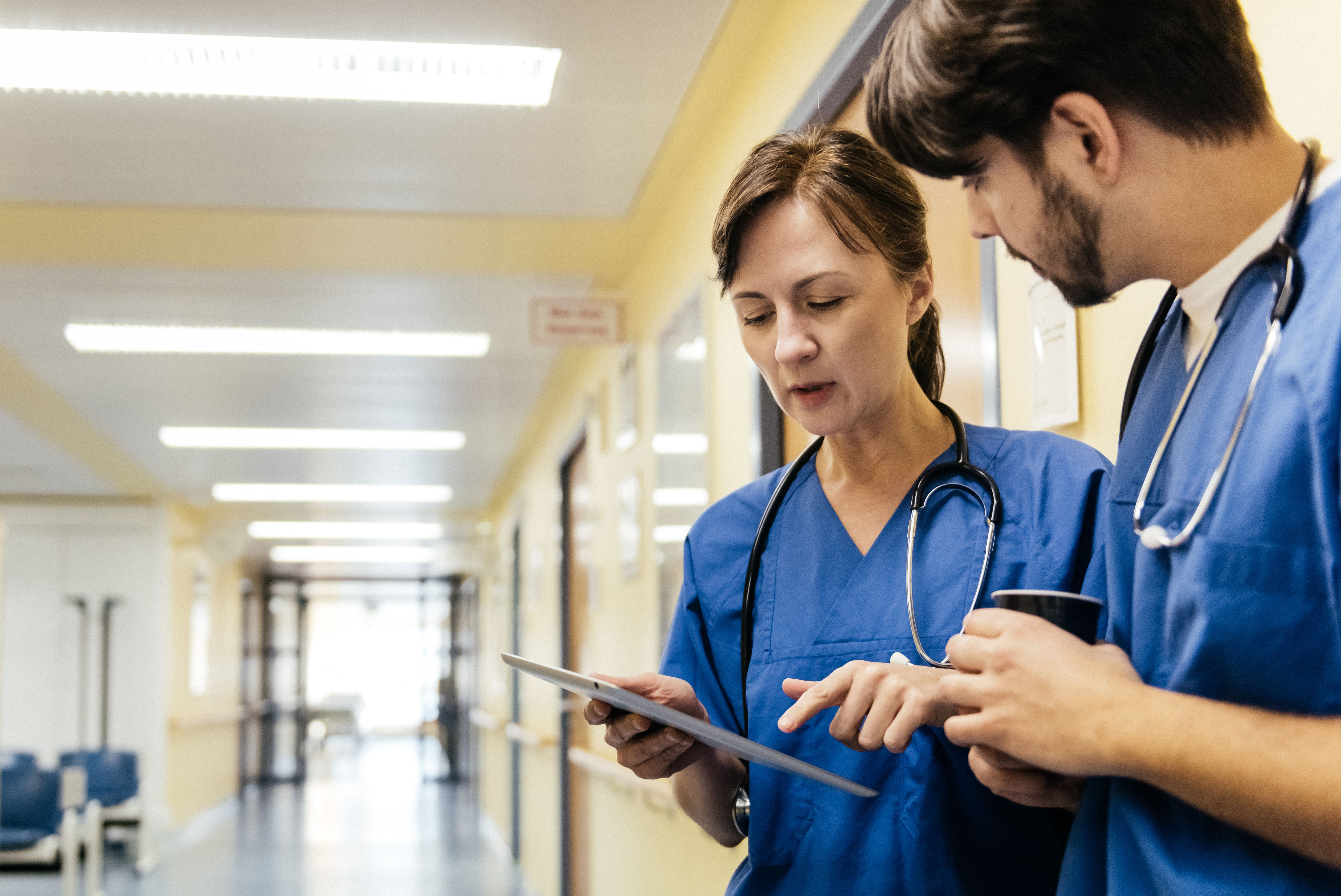 Mature white female doctor and her younger male colleague discussing patient record on a digital tablet while standing in a hospital corridor