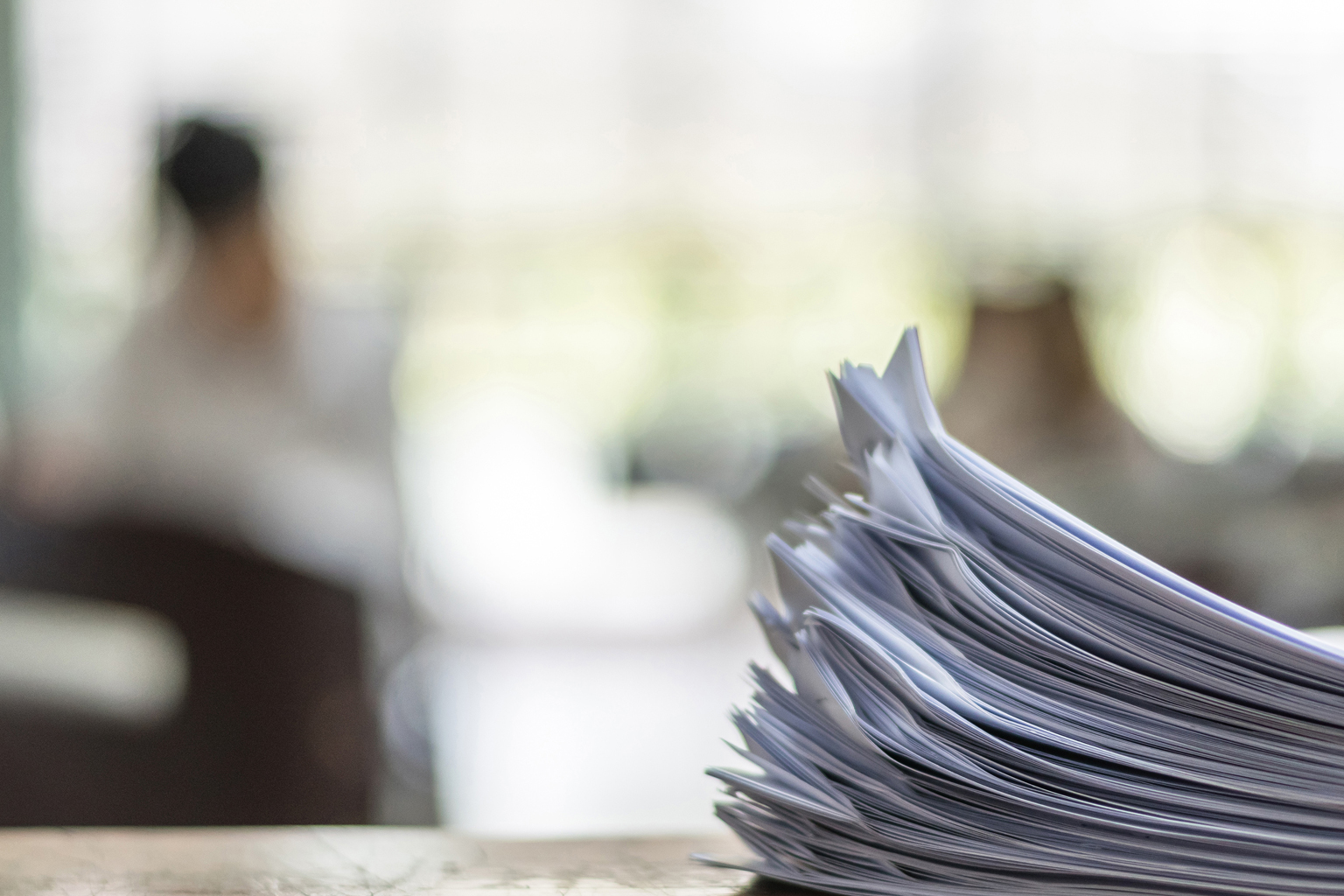 Stack of papers in foreground with two people in office blurred out in background