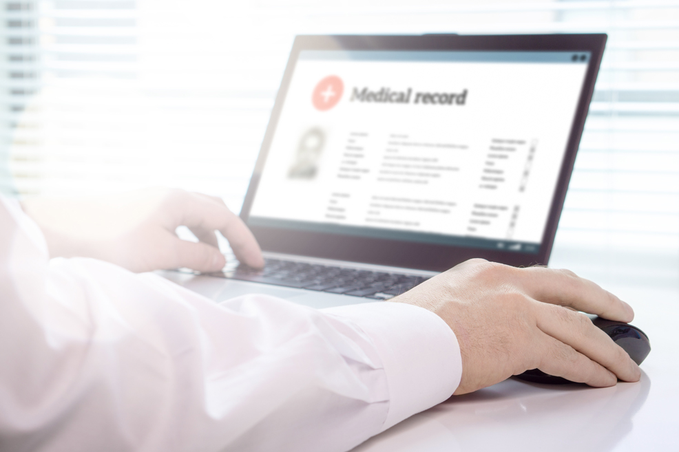 Close up on person's arms on laptop and mouse with medical record page open on the screen