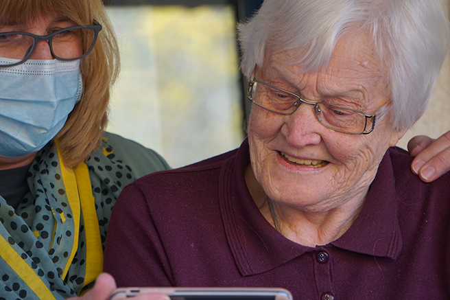 Masked woman sitting with senior woman holding mobile up while they watch the screen