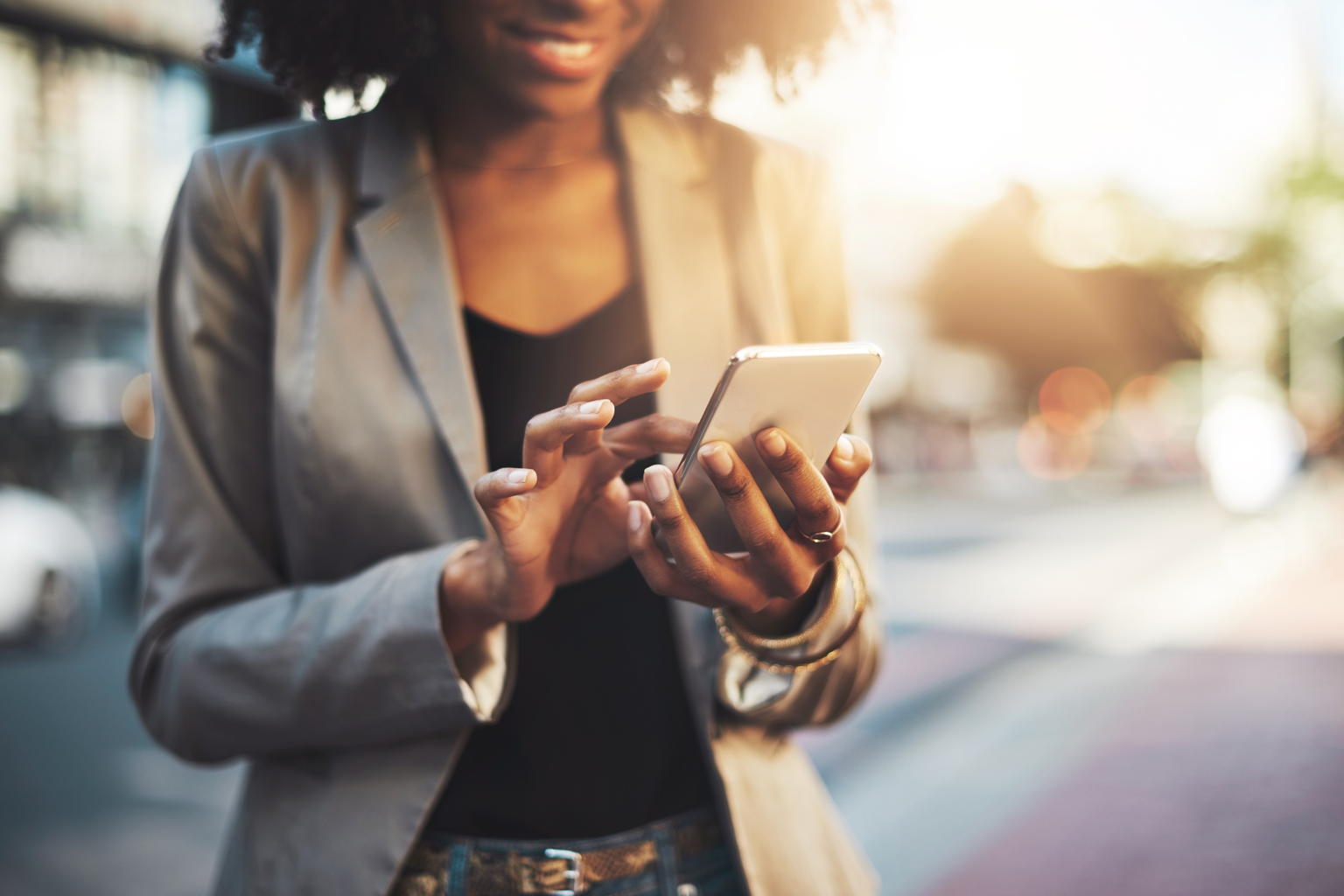 Smiling woman with curly hair and grey, gray blazer holding and using her phone outside, gold jewelry , Q3 2021, TAA NA US - Preparer