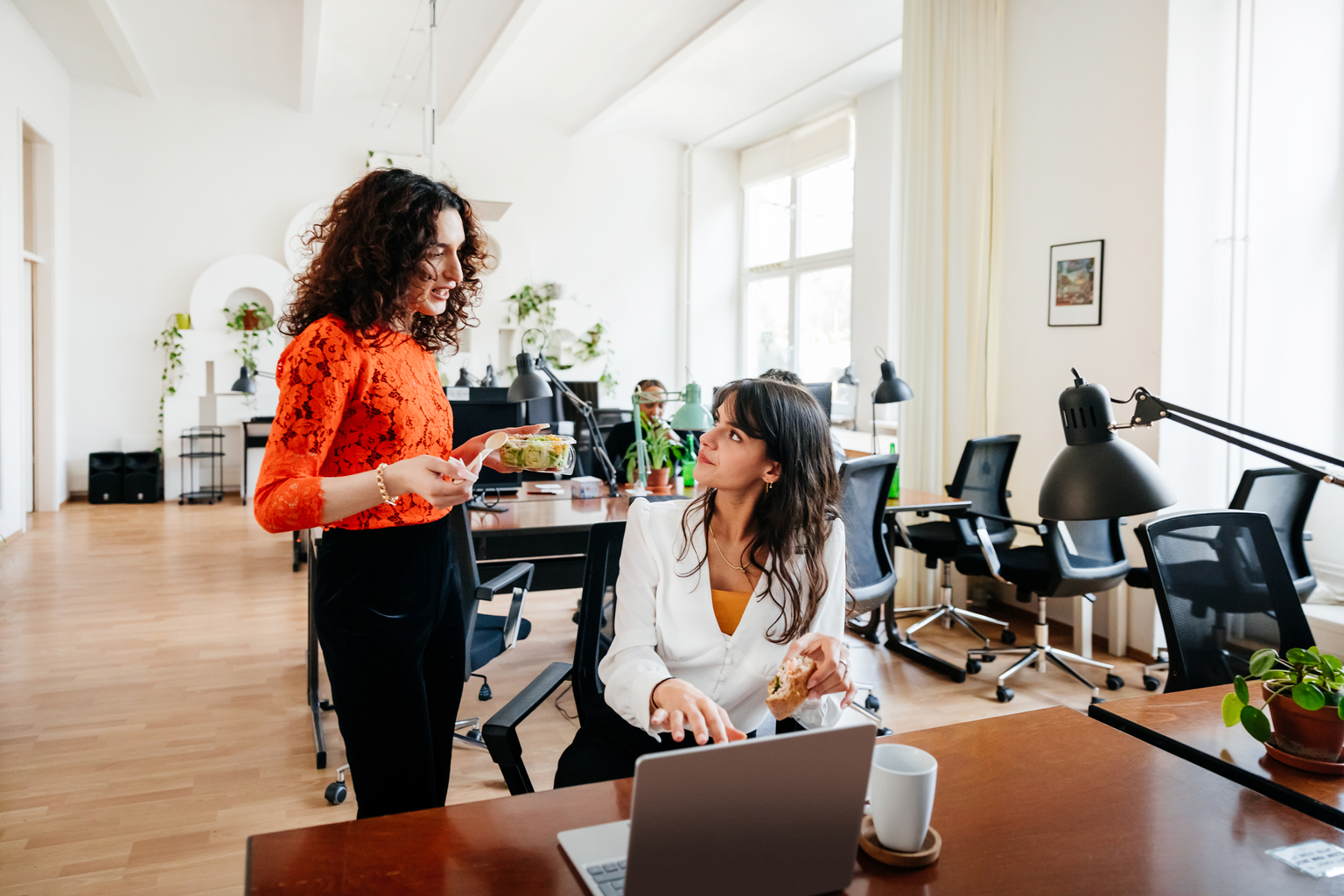 Two female coworkers eating lunch, discussing and working in the office