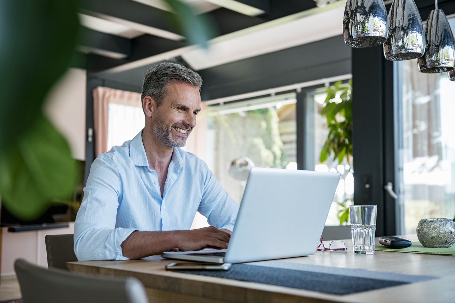 man with beard wearing blue collared dress shirt smiling at his macbook, laptop in at home office, tree focus in corner, silver ceiling lights, Q3 2021, TAA NA US - Preparer