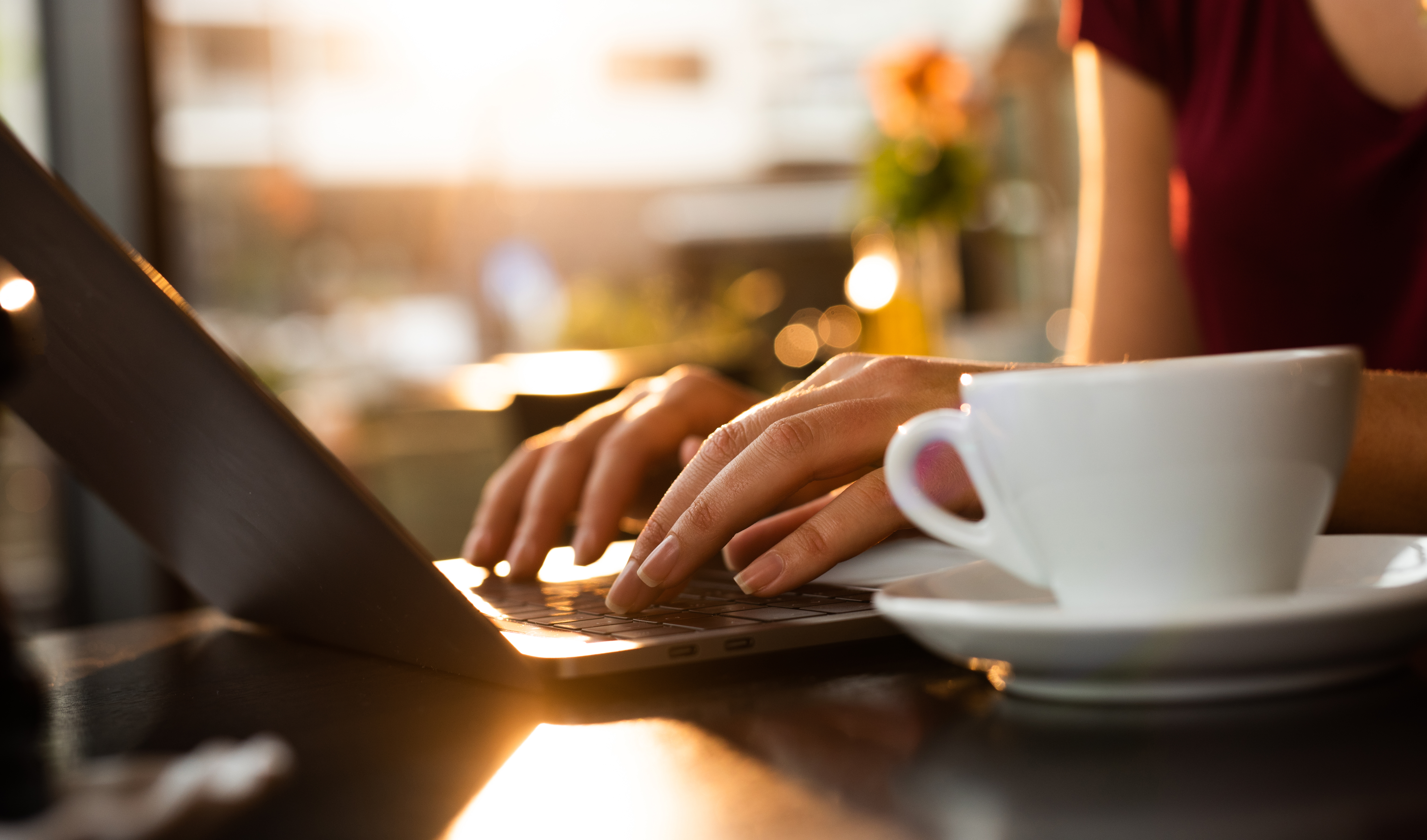 Woman with maroon t shirt typing on her macbook at a coffee shop with white mug, teacup, Q3 2021, TAA NA US - Preparer