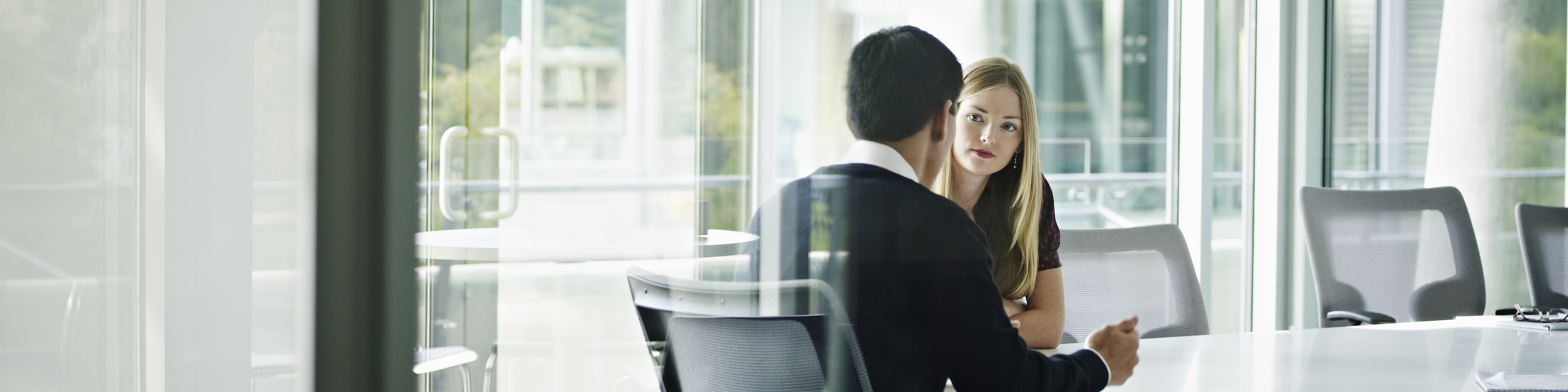 Businesswoman and businessman in discussion at table in glass walled conference room.