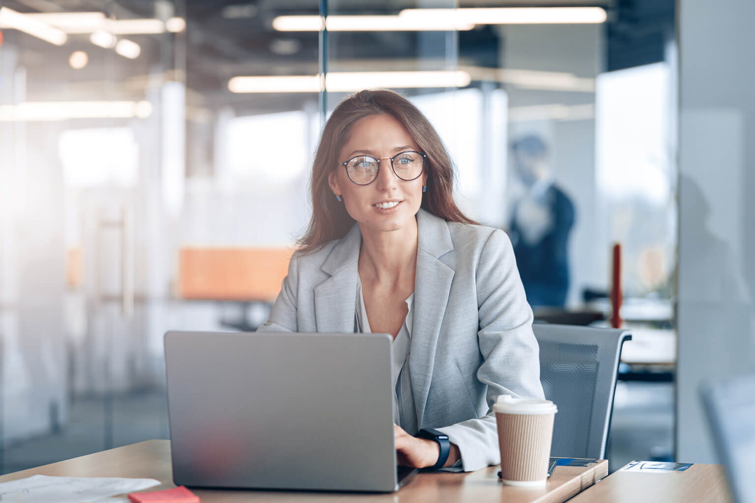 A woman sitting at a desk with a laptop and a cup of coffee, looking off into the distance