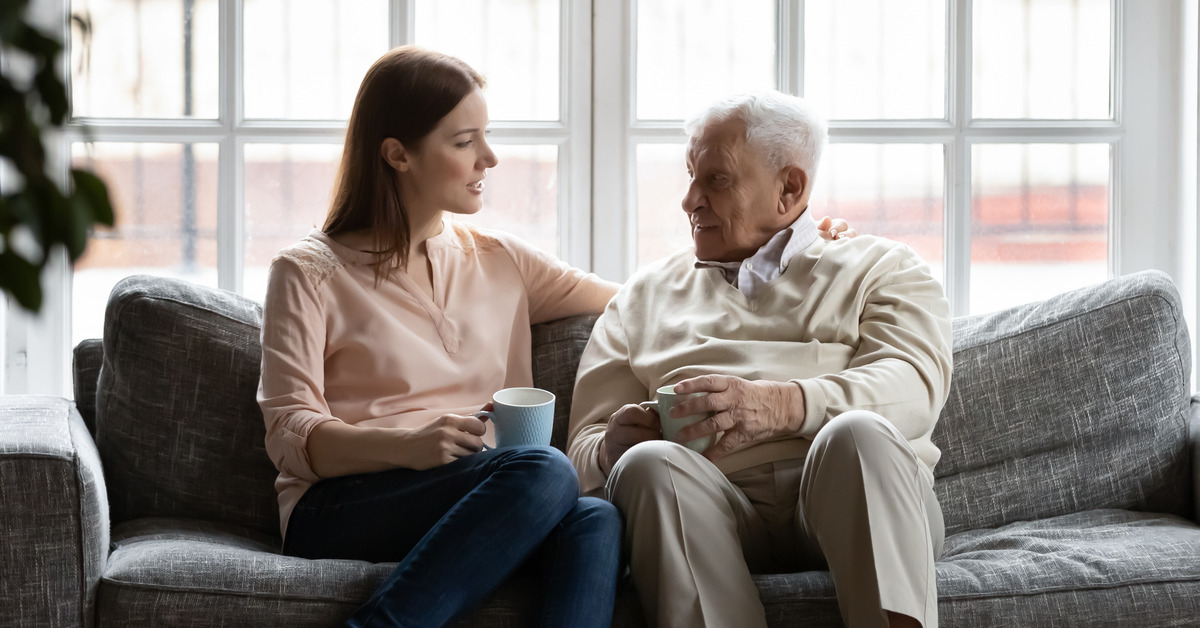 Happy elderly father sit on couch in living room talk with adult daughter drinking tea together