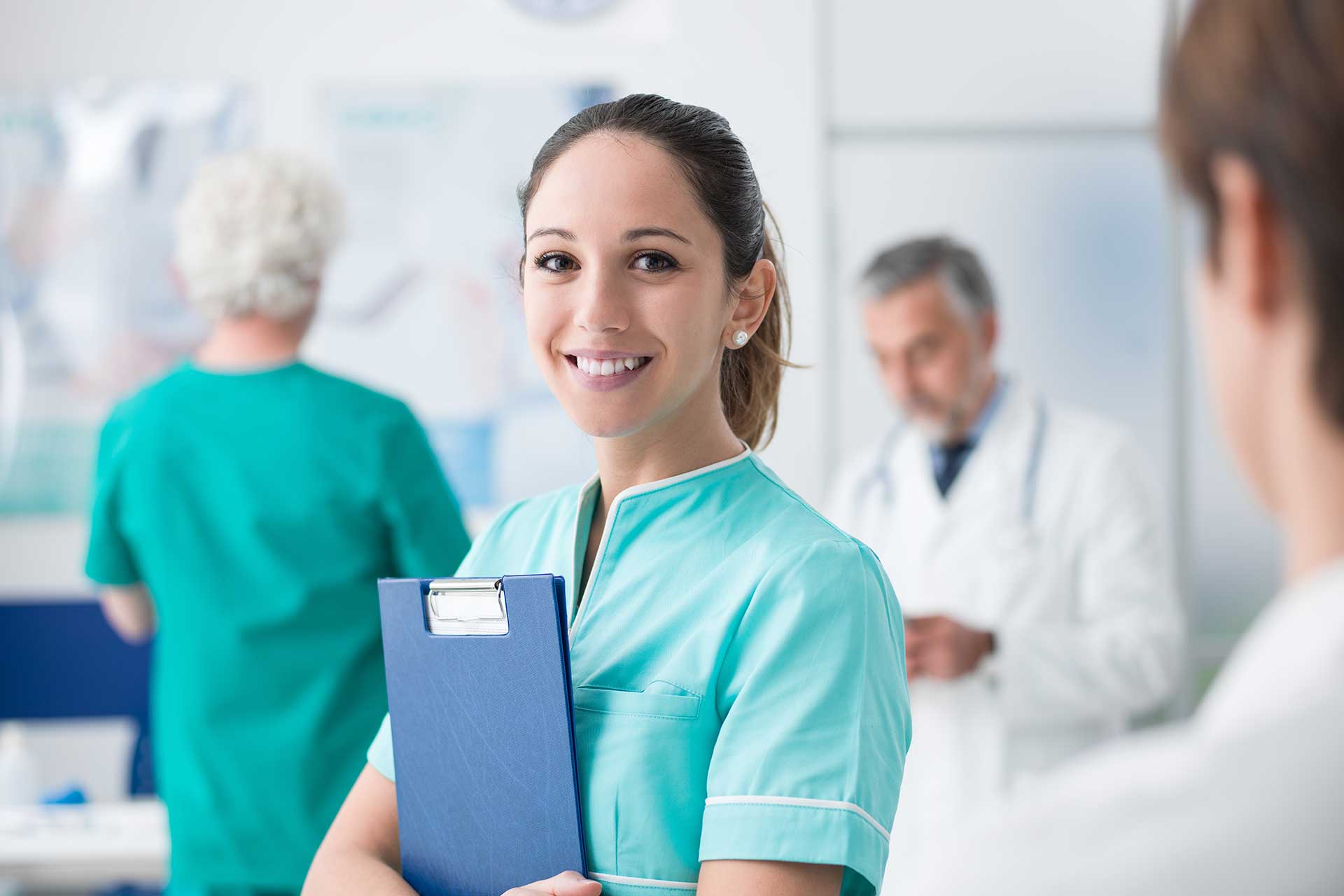 Nurse holding file to her chest and looking at camera in blurred background of hospital setting