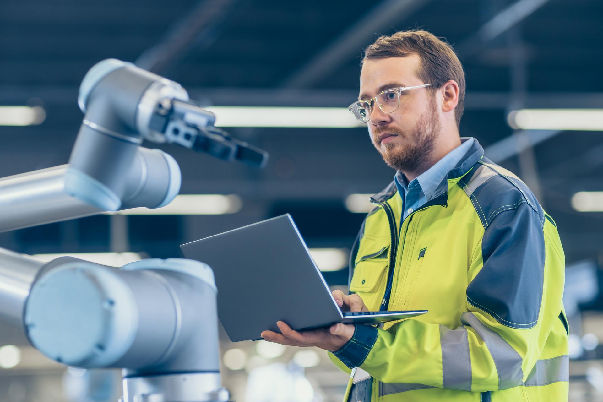 At the Factory: Automation Engineer Uses Laptop for Programming Robotic Arm. New Era in Automatic Manufacturing Industry