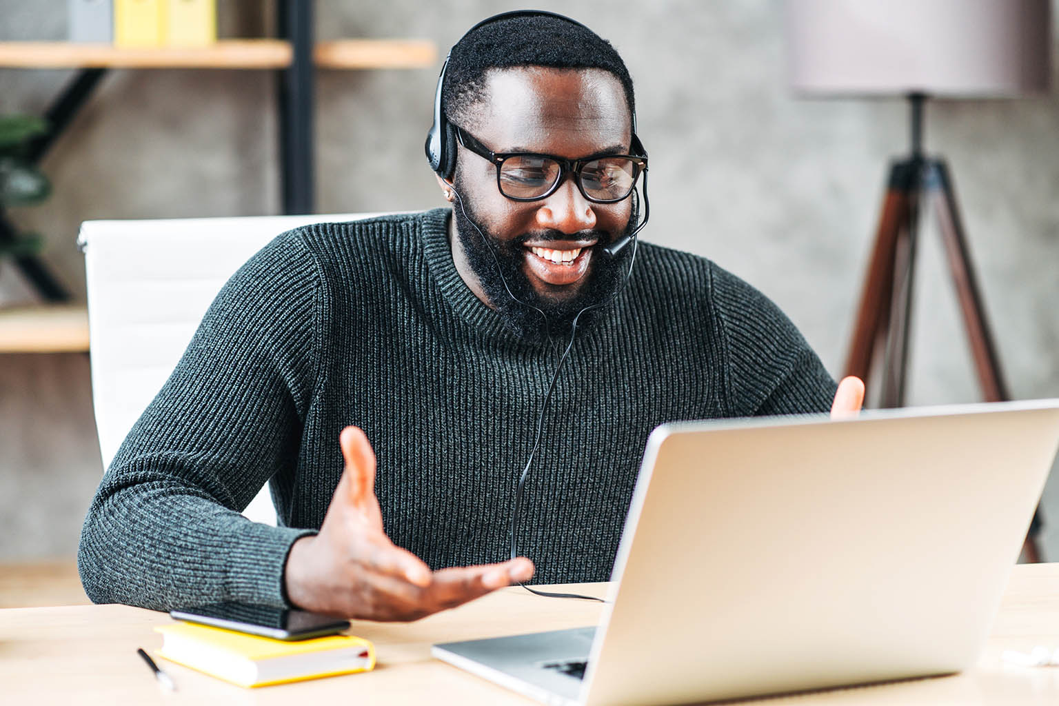 Happy man sitting at open laptop with headset on and talking to someone