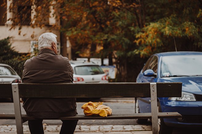 Older man facing away from camera, sitting on sidewalk bench