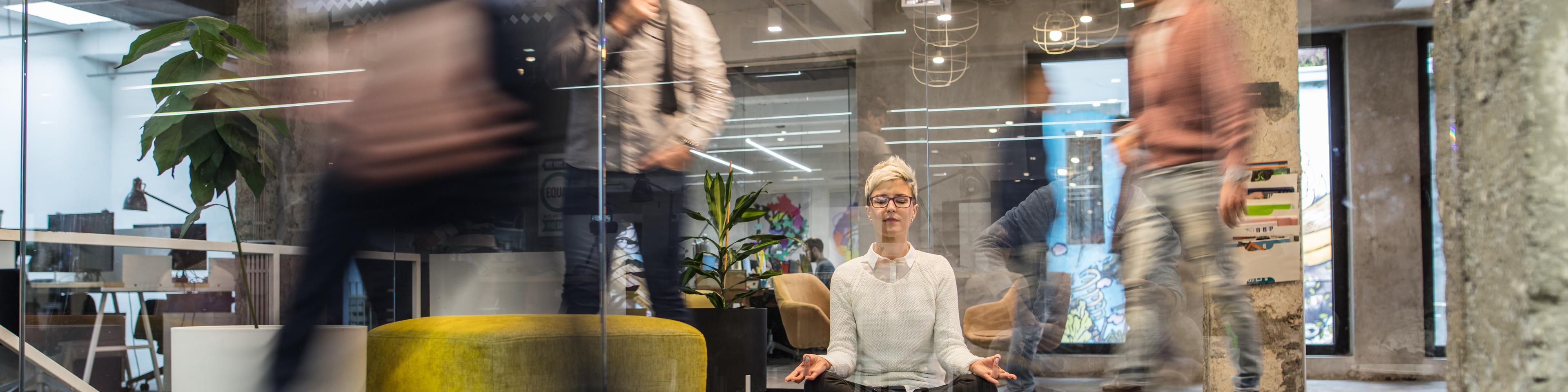 Young female entrepreneur meditating with eyes closed in busy office. 